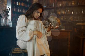 Scary demonic woman holds human skull, demons casting out. Exorcism, mystery paranormal ritual, dark religion, night horror, potions on shelf on background