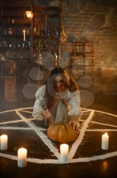 Crazy demonic woman with pumpkin sitting in magic circle with candles, demons casting out. Exorcism, mystery paranormal ritual, dark religion, night horror, potions on shelf on background