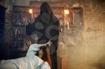 Male exorcist in black hood casting out devil from creepy woman. Exorcism, mystery paranormal ritual, dark religion, night horror, potions on shelf on background