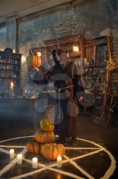 Male exorcist in black hood holds pumpkin near the magic circle. Exorcism, mystery paranormal ritual, dark religion, night horror, potions on shelf on background