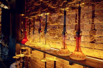 A row of hookahs with glass bulbs at the brick wall, nobody. Shisha bar equipment, traditional smoke culture, tobacco aroma