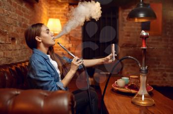 Young woman makes selfie with hookah in bar, mesmerizing cloud. Shisha smoking, traditional bong culture, tobacco aroma for relaxation, rest with hooka