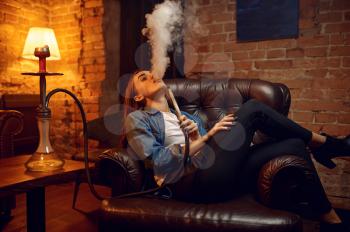 Young woman blows smoke in hookah bar, chill out. Shisha smoking, traditional bong culture, tobacco aroma for relaxation, rest with hooka