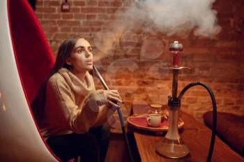 Young woman smokes hookah in bar, chill out. Shisha smoking, traditional bong culture, tobacco aroma for relaxation, rest with hooka
