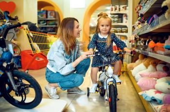 Mom and pretty little girl choosing bicycle in kid's store. Mother and adorable daughter near the showcase in toyshop, happy childhood, family makes a purchase in a shop
