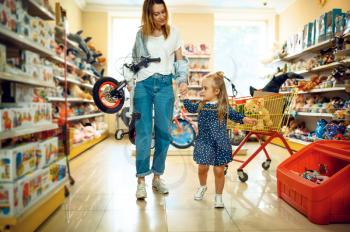 Mother and pretty little girl buying bicycle in kid's store. Mom and adorable daughter near the showcase in toyshop, happy childhood, family makes a purchase in shop