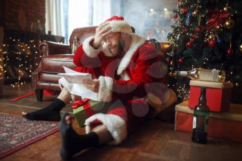 Bad drunk Santa claus reads letters under christmas tree, nasty party, humor. Unhealthy lifestyle, bearded man in holiday costume, new year and alcoholism