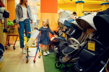 Mother and pretty little girl buying baby stroller in store. Mom and adorable daughter near the showcase in children's shop, happy childhood, family makes a purchase in market