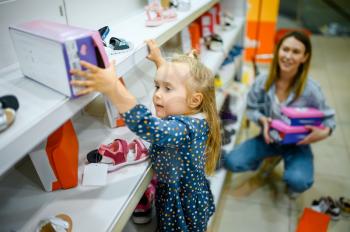 Little baby climbs on showcase to takes a shoes in kid's store. Mom and adorable girl buying sandals in children's shop, happy childhood, family makes a purchase in market