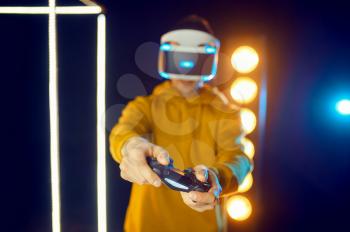 Man plays the game using virtual reality helmet and gamepad in luminous cube, front view. Dark playing club interior, spotlight on background, VR technology with 3D vision