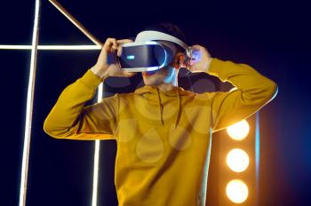 Young man plays the game using virtual reality headset and gamepad in luminous cube. Dark playing club interior, spotlight on background, VR technology with 3D vision