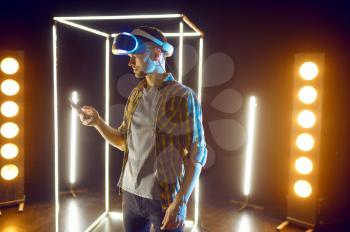 Man gaming in virtual reality headset and gamepad in luminous cube. Dark playing club interior, spotlight on background, VR technology, 3D vision