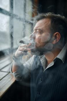 Man at the windowsill and smokes a cigar, vintage office interior on background. Tobacco smoking culture, specific flavor. Male smoker looks at the window