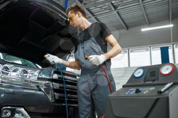 Male mechanic refills the air conditioner, car service. Vehicle repairing garage, man in uniform, automobile station interior on background