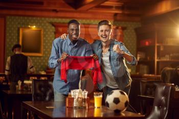 Two male football fans with red scarf and ball watching game tv broadcast, friends in bar. Group of people relax in pub, night lifestyle, friendship, sport victory celebration