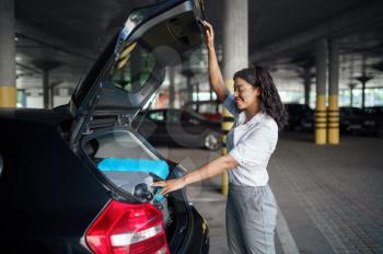Young woman puts suitcases into the car on parking. Female traveler with luggage in vehicle park lot, passenger with many bags. Girl with baggage near automobile