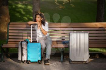 Young woman with suitcase sitting on the bench in summer park. Female traveler with luggage leisures outdoors, passenger with bag resting in nature. Girl with baggage relax on city alley