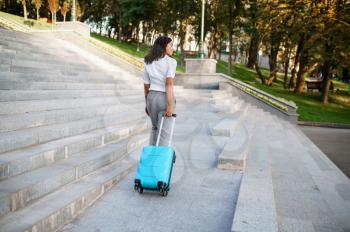 Happy woman poses with suitcase on the stairs in summer park. Female traveler with luggage leisures outdoors, passenger with bag resting in nature. Girl with baggage relax on city alley
