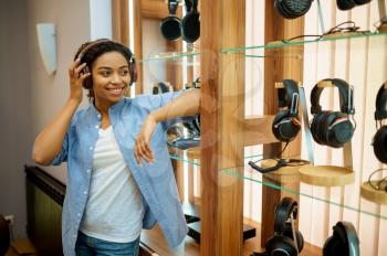 Young woman listening to music in headphones store. Female person in audio shop, showcase with earphones on background, buyer in the multimedia salon