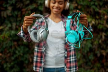 Music fan in headphones listening to music in summer park. Female audiophile walking outdoors, girl in earphones, green bushes on background