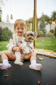 Kid play with funny dog on trampoline in the garden. Child with puppy leisures on backyard. Little girl and her pet having fun on playground outdoors, happy childhood