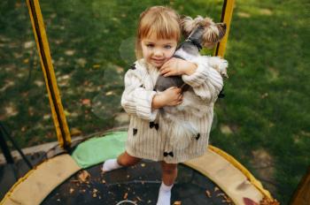Kid hugs funny dog on trampoline in the garden. Child with puppy leisures on backyard. Little girl and her pet having fun on playground outdoors, happy childhood