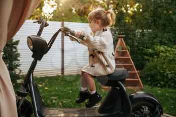Little girl sitting on vintage scooter in the garden. Female child poses on motor vehicle on backyard. Kid having fun on playground outdoors, happy childhood