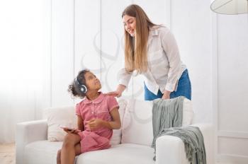 Mother and her little daughter in headphones on sofa in living room.Mom and female child leisures in their house together, good relationship, parental care