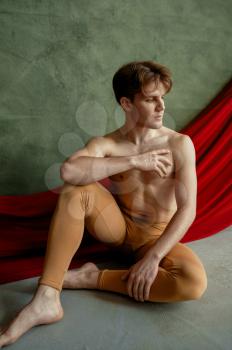 Male ballet dancer, dancing studio, grunge wall and red cloth on background. Performer with muscular body, grace and elegance of movements