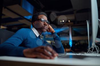 Man in glasses works on computer, office lifestyle. Male person at desktop, dark interior on background, modern workplace