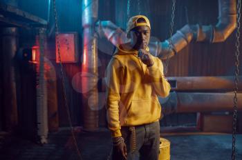 Stylish rapper in yellow hoodie and baseball cap poses in studio with cool underground decoration. Hip-hop performer, rap singer, break-dance