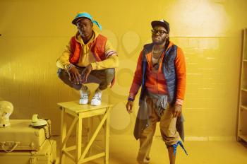Two stylish rappers with gold jewelry in cool studio, yellow background. Hip-hop performers, trendy rap singers, break-dancers