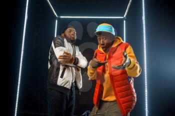 Two stylish rappers poses in glowing cube, studio with dark background. Hip-hop performers, break-dancers
