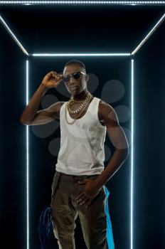 Serious rapper in gold jewelry poses in illuminated cube, dark background. Hip-hop performer, rap singer, break-dance performing, entertainment lifestyle, breakdancer