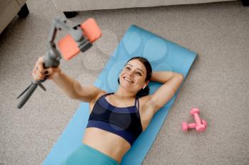 Slim woman lying on mat, online fit training with phone. Female person in sportswear, internet sport workout, room interior on background