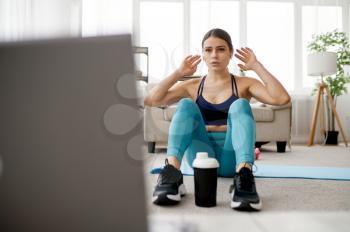 Slim woman sits on mat, online fitness training at the laptop. Female person in sportswear, internet sport workout, room interior on background