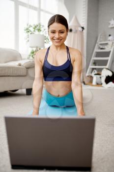 Smiling girl sits on the floor at home, online fit training at the laptop. Female person in sportswear, internet sport workout, room interior