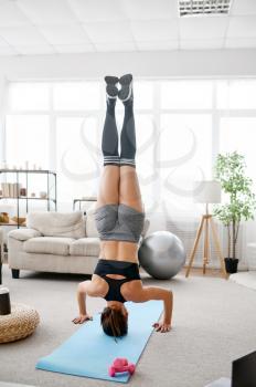 Woman stands upside down, online yoga training at the laptop. Female person in sportswear, internet sport workout, room interior on background