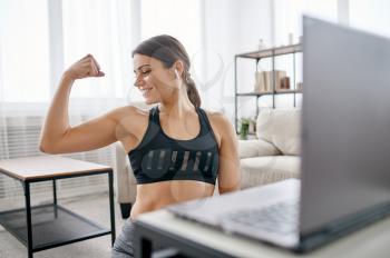 Woman shows her muscles, online fitness training at the laptop. Female person in sportswear, internet sport workout, room interior on background