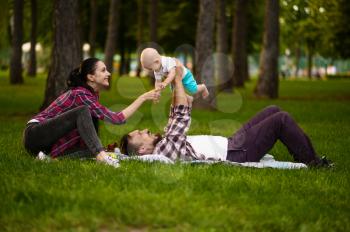 Cheerful parents and little baby play on grass in summer park. Mom and dad with male kid on lawn, picnic with child on plaid in the forest, family happiness