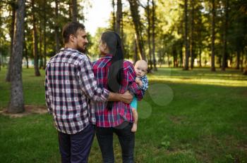 Mother and father walks together with little baby in summer park. Mom and dad with male kid on lawn, parents on picnic with child in the forest, family happiness