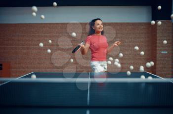 Woman among flying ping pong balls, table tennis training in gym, female player. Sportive girl playing table-tennis indoors, sport game with racket, active healthy lifestyle