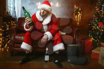 Bad Santa claus celebrate christmas with bottle of alcohol on couch. Unhealthy lifestyle, bearded man in holiday costume, new year and alcoholism