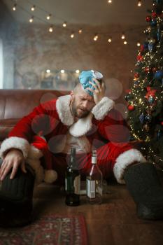 Vile Santa claus with hangover sitting under christmas tree with bottle of alcohol, nasty party, humor. Unhealthy lifestyle, bearded man in holiday costume, new year and alcoholism