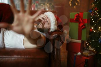 Bad Santa claus with hangover lying on sofa, nasty party, humor. Unhealthy lifestyle, bearded man in holiday costume, new year and alcoholism