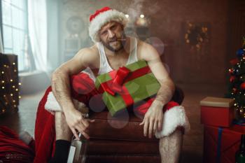 Bad Santa claus with cigar and bottle of alcohol sitting on couch, nasty party, humor. Unhealthy lifestyle, bearded man in holiday costume, new year and alcoholism
