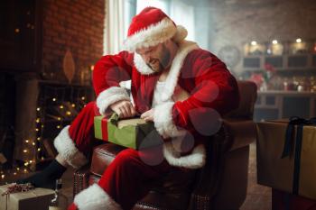 Bad drunk Santa claus opens gifts under christmas tree, nasty party, humor. Unhealthy lifestyle, bearded man in holiday costume, new year and alcoholism