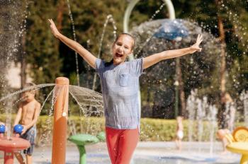 Little funny girl in splashes on water playground in summer park. Child leisures in aquapark, aquatic adventure, female kid having fun in fountain, waterpark