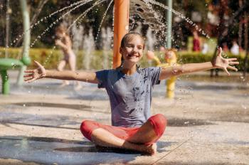 Happy little girl sitting in yoga pose on water playground in summer park. Child leisures in aquapark, aquatic adventure, female kid having fun in fountain, waterpark