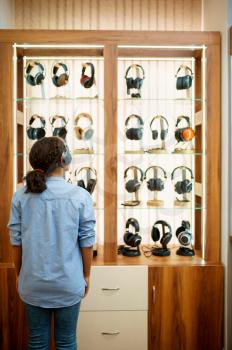 Woman choosing headphones in acoustics store, back view. Female person in audio shop, showcase with earphones on background, buyer in multimedia salon